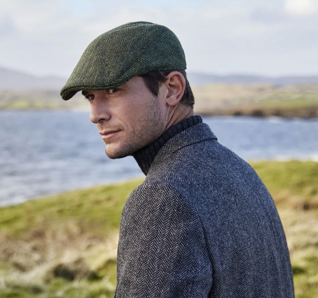 Man wearing a green cap and blue coat standing by a body of water with grassy hills in the background.